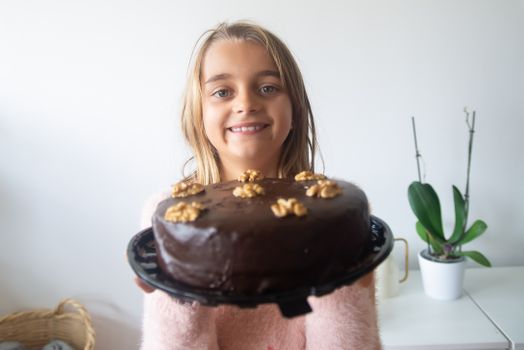 A happy girl proudly holds a homemade chocolate cake topped with walnuts indoors.