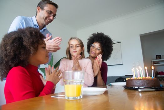 A diverse family clapping and celebrating a birthday with a chocolate cake and candles indoors.