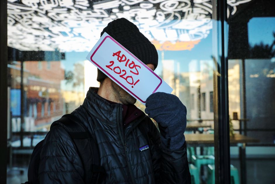 Person in warm jacket holding 'Adios 2020' sign in an urban setting. Conceptual farewell theme.