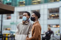 Serious African American gays wearing protective masks standing on street with residential building on blurred background in city during coronavirus pandemic