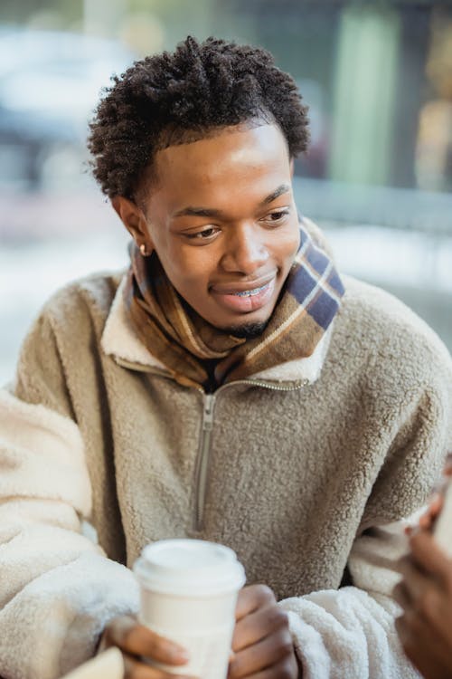 Positive African American male in warm clothes with paper cup of hot beverage sitting on terrace of cafe on blurred background