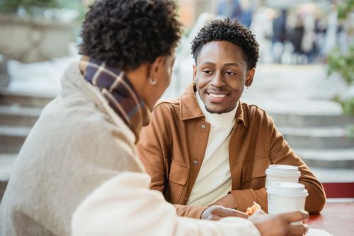 Free Happy African American male with faceless partner sitting at table with food and paper cups of hot beverage while having break on terrace of cafe on blurred background Stock Photo