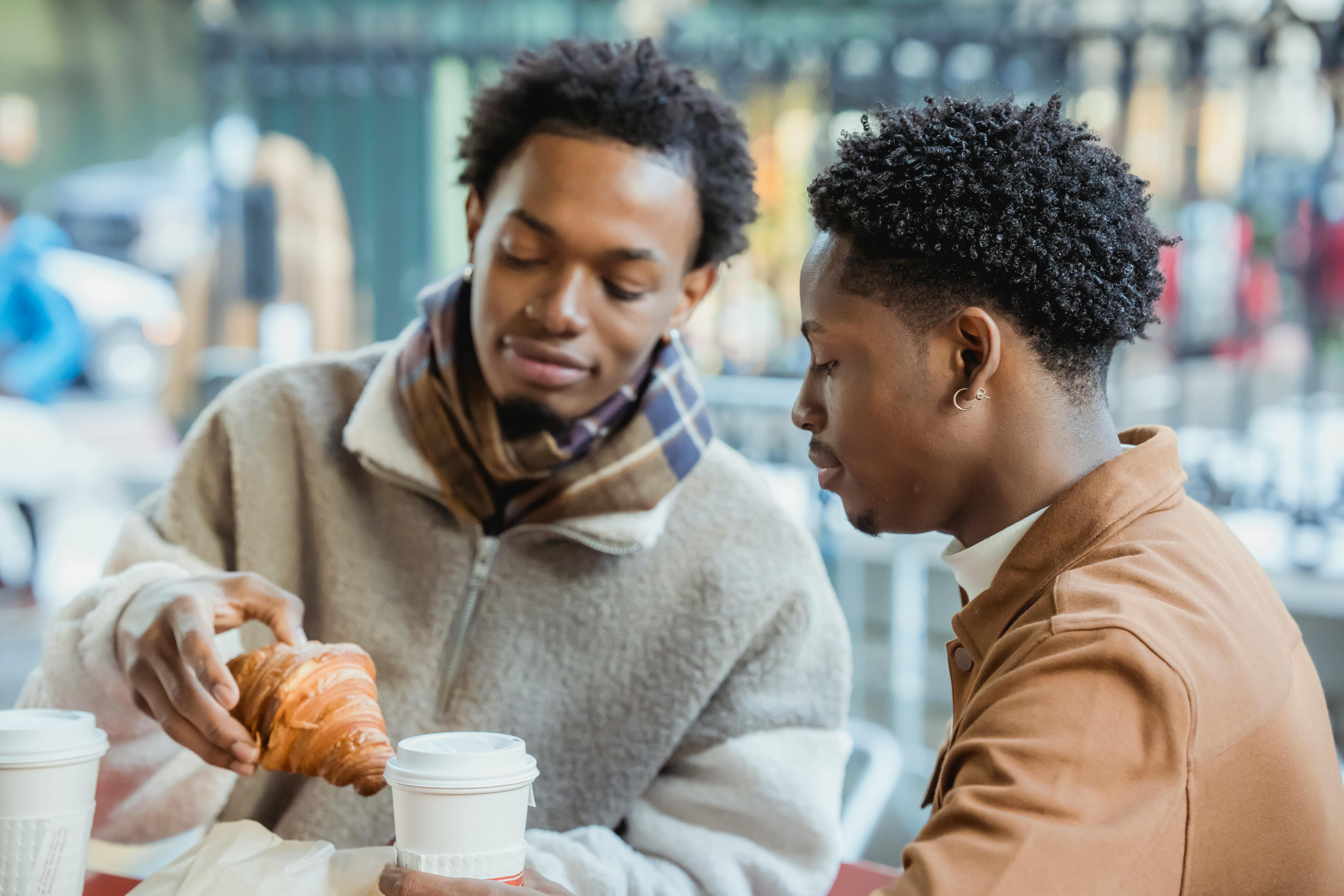 Content African American homosexual couple sitting at table with paper cups of coffee and fresh pastry while having lunch on terrace of cafeteria