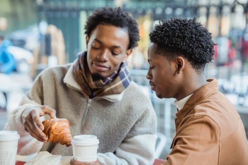Free Content African American homosexual couple sitting at table with paper cups of coffee and fresh pastry while having lunch on terrace of cafeteria Stock Photo