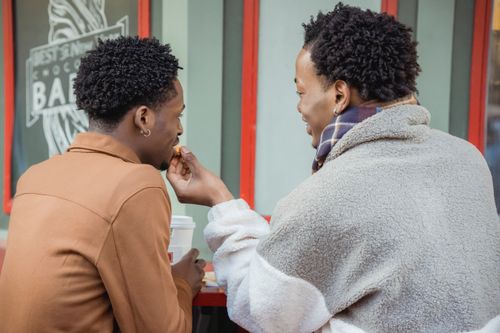 Free Side view of African American gay feeding black partner while having lunch at table with paper cup of coffee in cafeteria Stock Photo