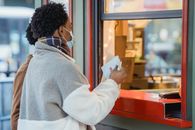 Side view of African American male couple in protective masks standing near window of store while ordering food on street on blurred background