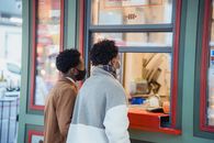 Side view of African American male in protective masks buying food while standing near window of store on street in city