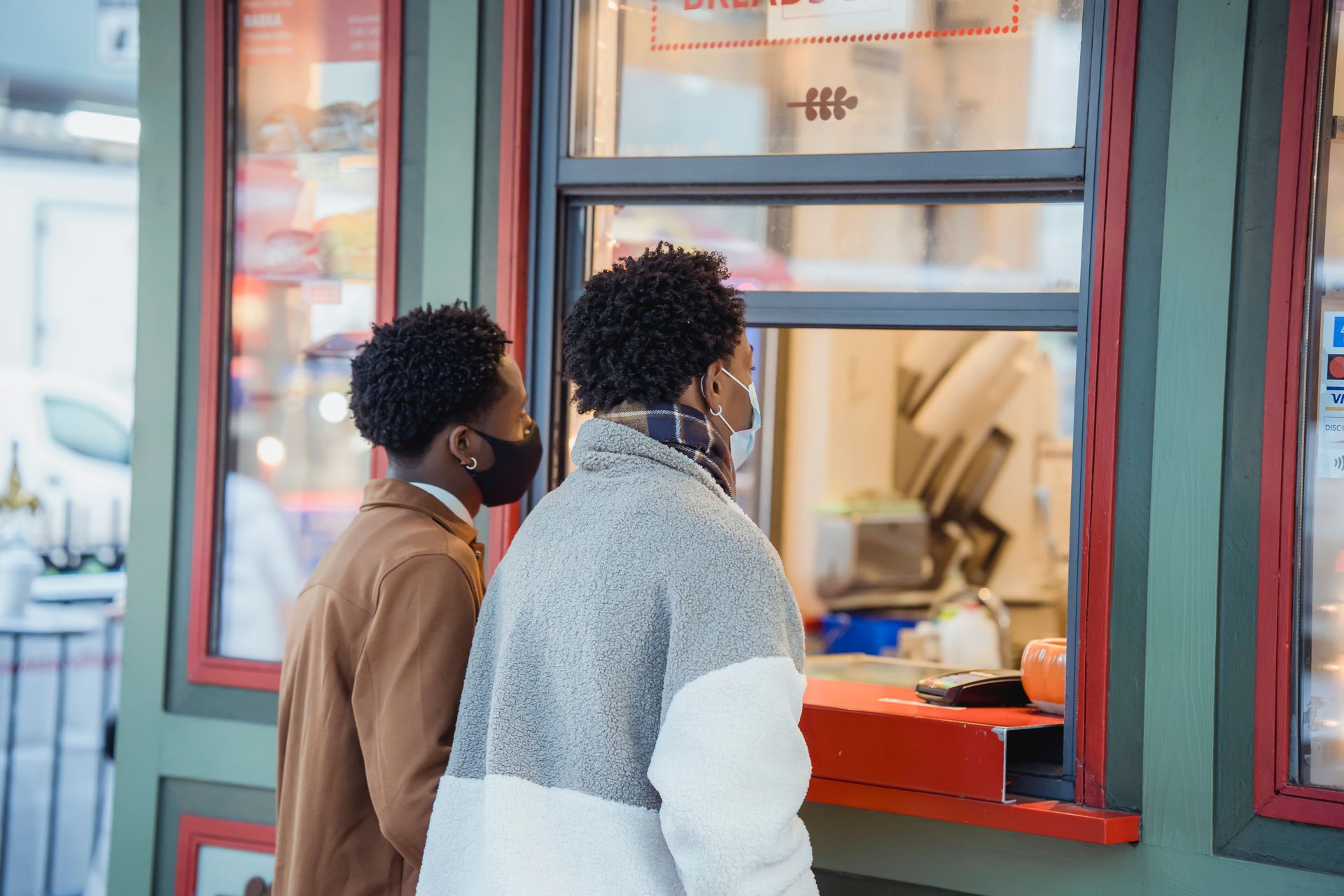 Side view of African American male in protective masks buying food while standing near window of store on street in city