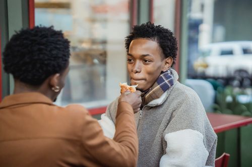 Free Anonymous man feeding cheerful African American boyfriend with tasty croissant while sitting on terrace of cafe in city on blurred background Stock Photo