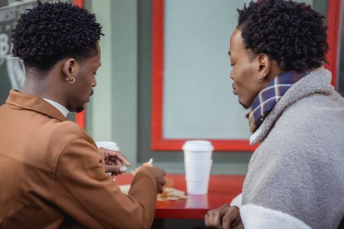 Free Side view of content African American homosexual couple sitting at table with food and paper cups of coffee while having lunch on terrace of cafe Stock Photo