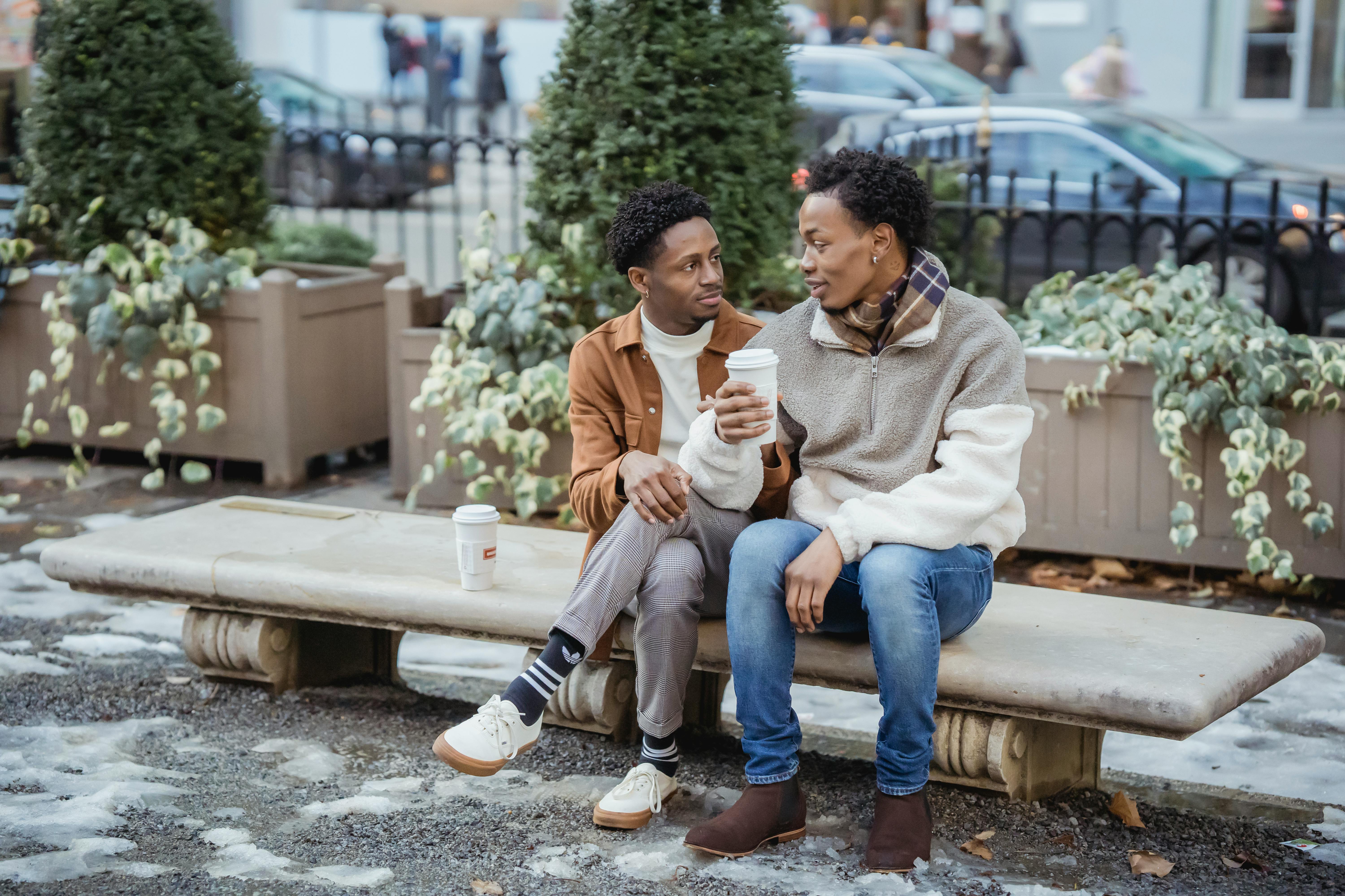 Full body of African American male couple with takeaway coffee looking at each other while sitting on bench on snowy street