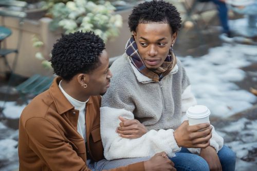 Free From above of tender African American homosexual couple with takeaway coffee looking at each other while sitting on snowy street on blurred background Stock Photo