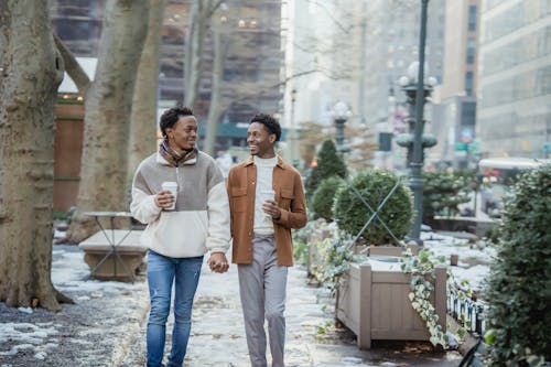 Cheerful African American homosexual couple with takeaway coffee holding hands and looking at each other while strolling on walkway on winter day