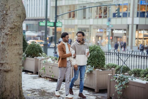Full body of positive African American male couple with takeaway hot beverages looking at each other while strolling on snowy walkway in city with buildings