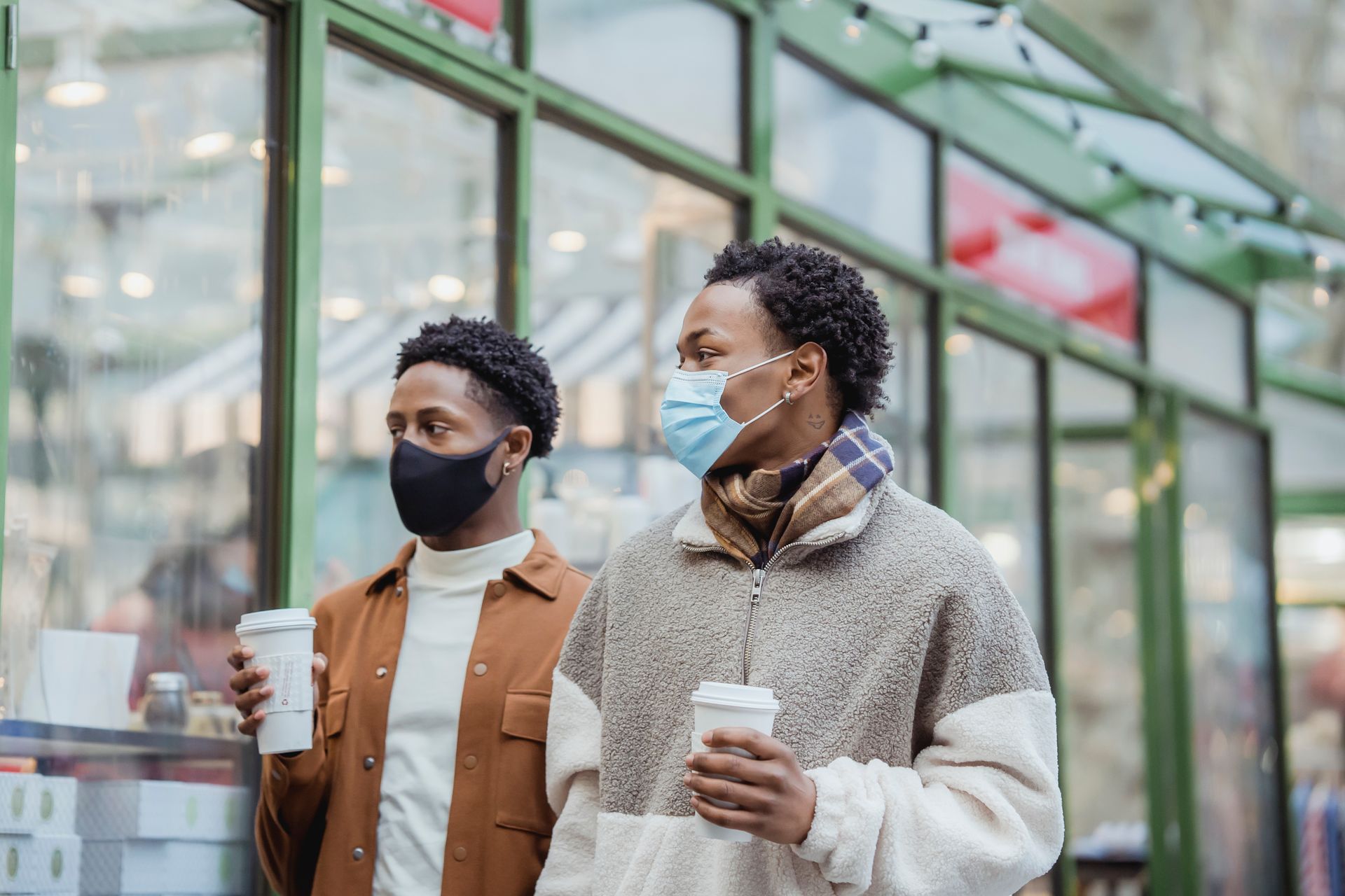 African American male couple in protective masks walking on street along building with paper cups of takeaway beverages in city