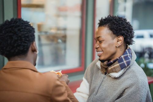 Free Smiling African American gay with anonymous partner eating tasty croissant while sitting on terrace of cafeteria on street on blurred background Stock Photo