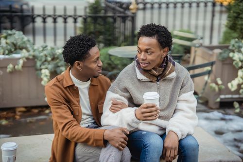 Free Positive African American homosexual couple with takeaway coffee looking at each other while sitting on bench during date on street Stock Photo