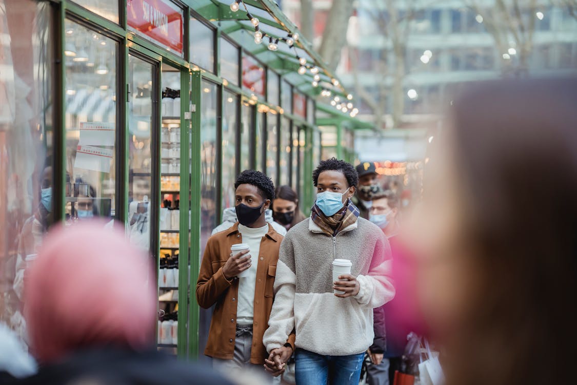 Free African American homosexual male in protective masks holding hands while strolling on street with takeaway coffee on street with people Stock Photo