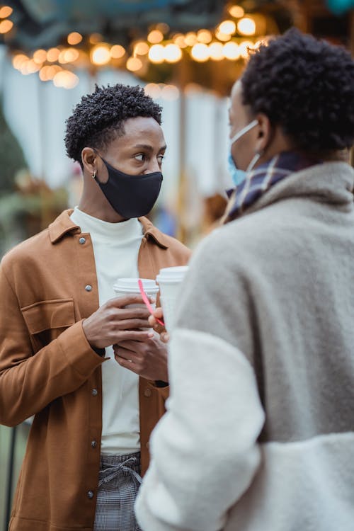 Unrecognizable young African American male friends in stylish clothes and protective masks standing on city street with takeaway coffee and communicating