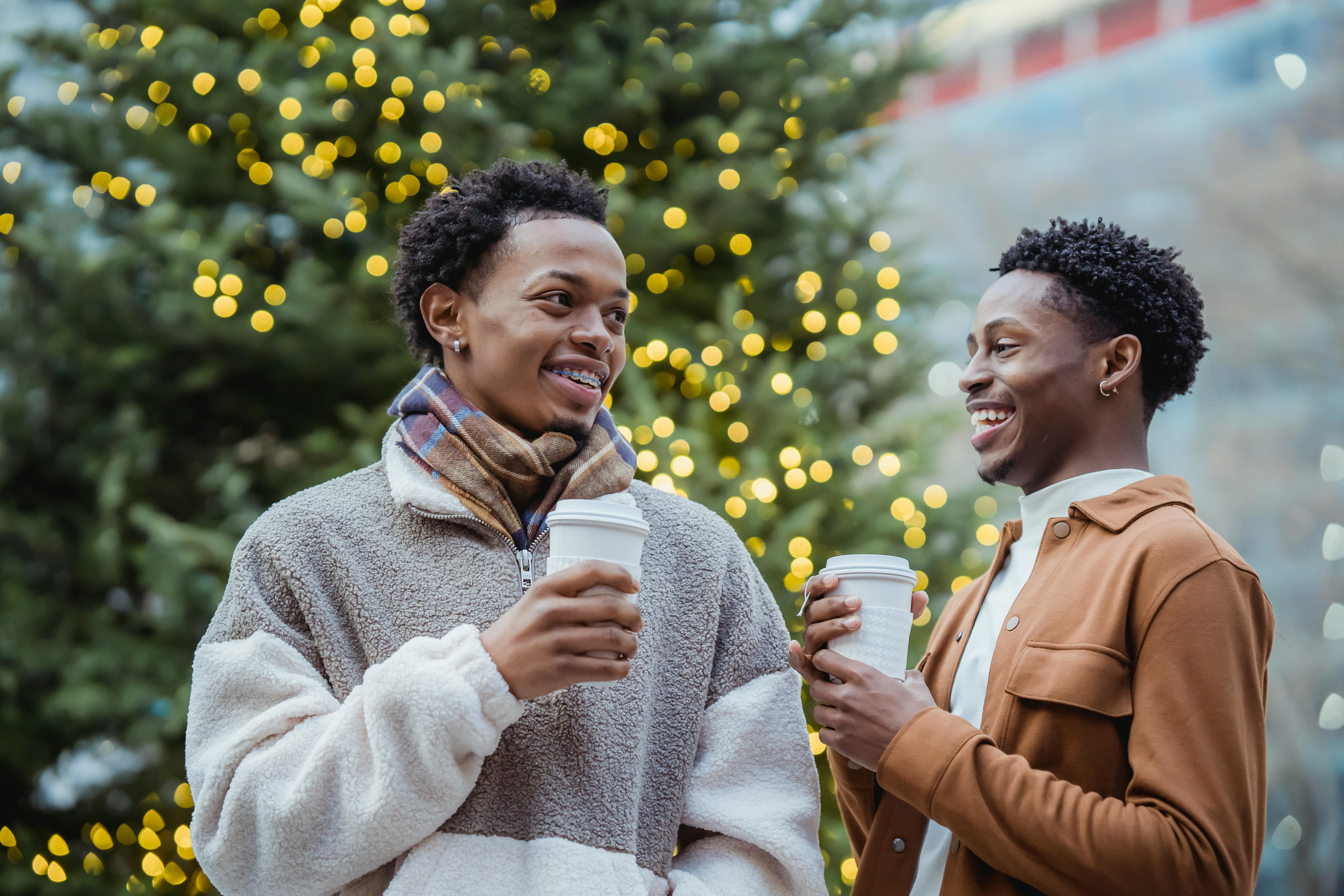 laughing young ethnic male friends drinking takeaway beverages near christmas tree on street