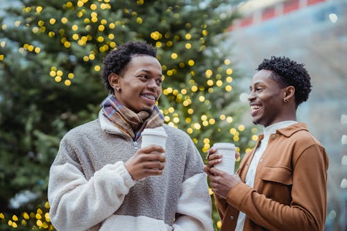 Free Laughing young ethnic male friends drinking takeaway beverages near Christmas tree on street Stock Photo