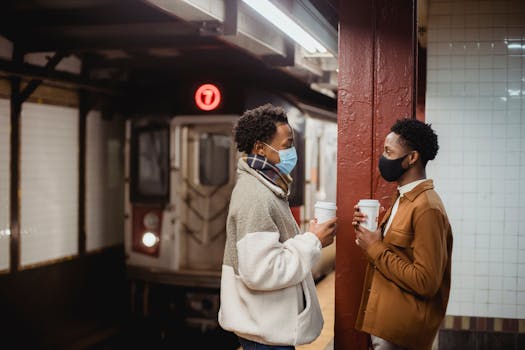 Two young men wearing masks chat while holding coffee in a subway station.