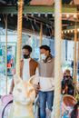 Anonymous black loving gay couple in trendy outfits and medical masks holding hands while standing on merry go round in amusement park