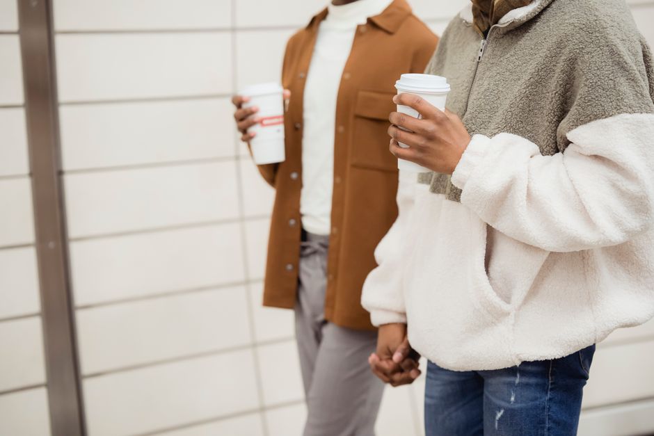 Crop anonymous African American homosexual couple wearing casual clothes with paper cups of hot beverage strolling together in subway