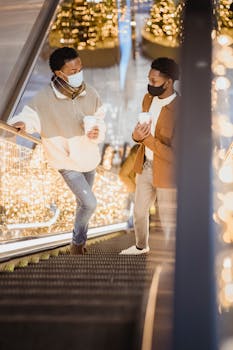 From above of young African American couple wearing trendy clothes and protective masks and talking while standing on escalator
