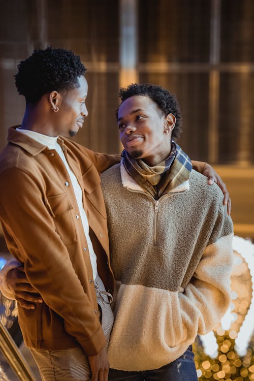 Positive African American male couple hugging while standing in street near building with windows and garlands in evening while looking at each other