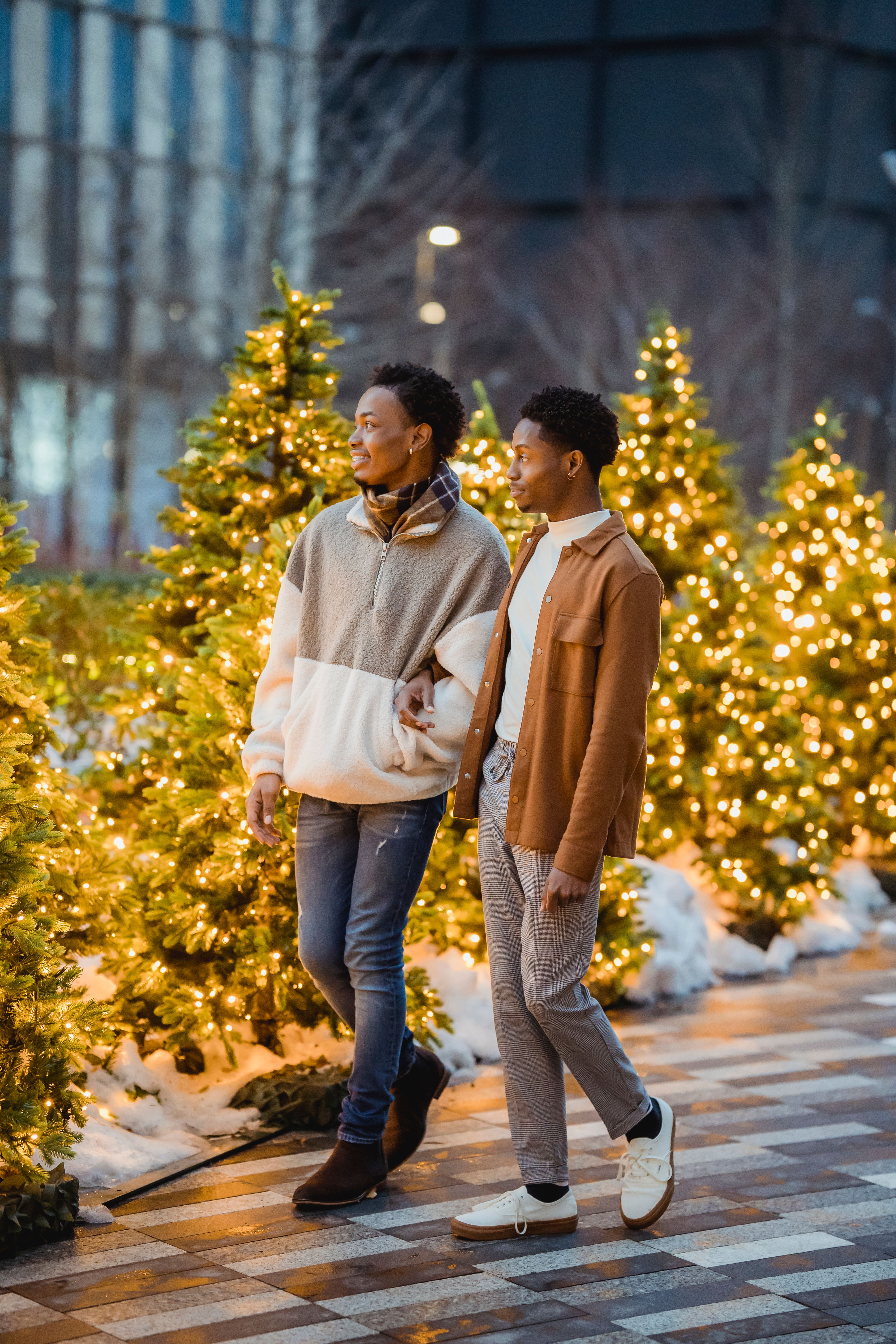 Full body of positive black gay couple strolling on pavement in city street near Christmas trees in snow decorated with garlands near buildings in evening