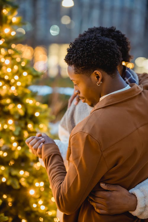 African American male couple in warm clothes embracing and holding hands while standing in town street near Christmas trees with glowing garlands in evening near building