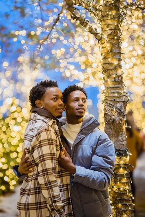 Black gay couple embracing while standing near trees with garlands