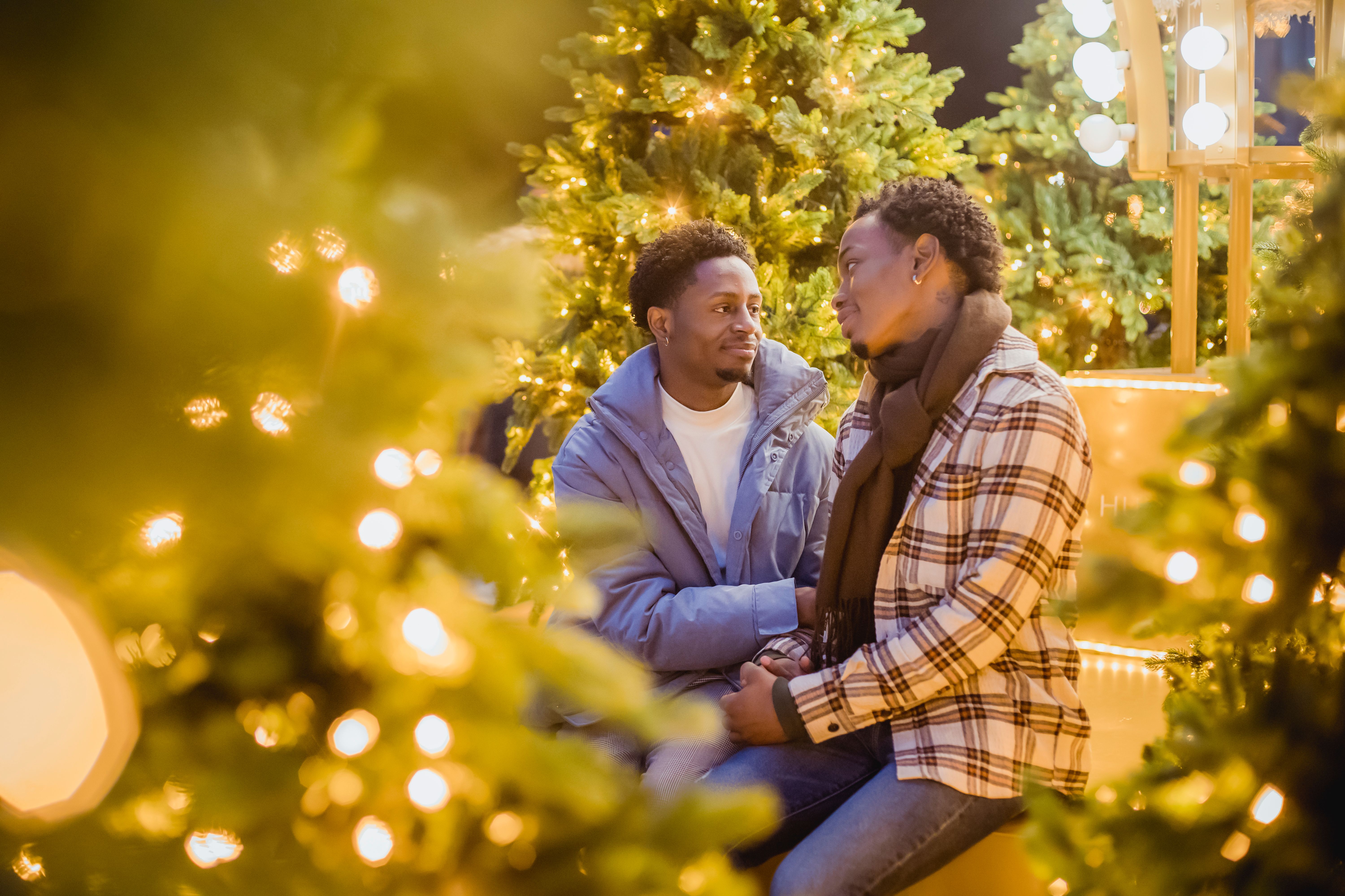 Content African American male gays looking at each other while sitting on street among green trees with glowing garlands during Christmas holidays