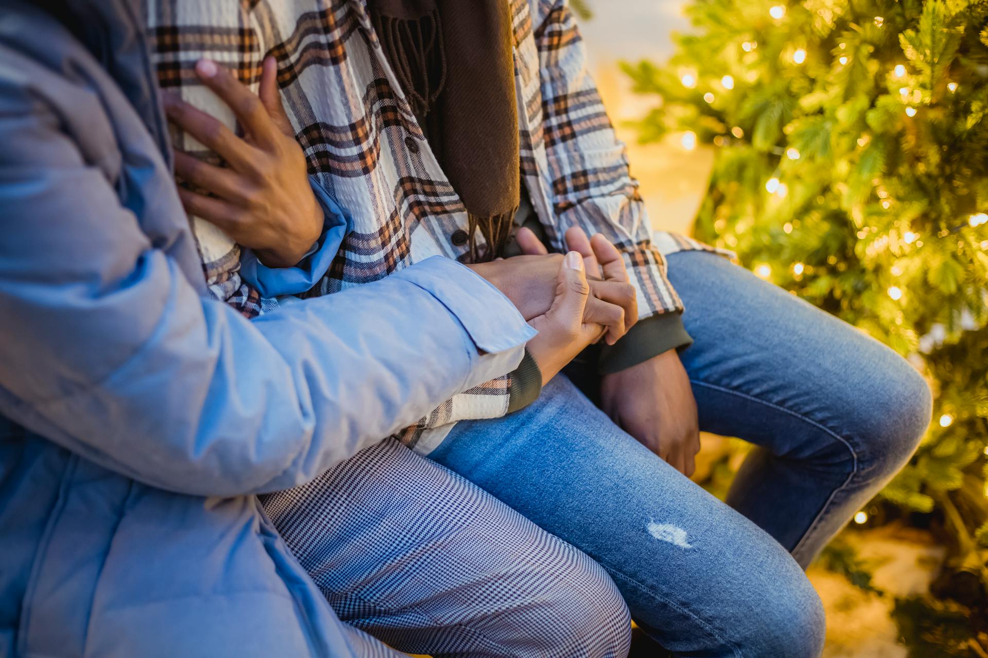 Unrecognizable romantic couple in holding hands and cuddling while sitting on street near Christmas tree with glowing lights during romantic date