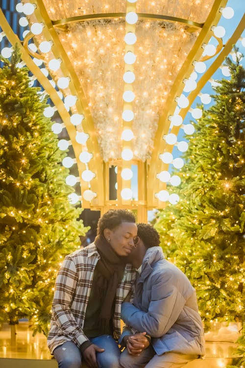 Loving African American homosexual couple embracing while sitting on street near decorations and Christmas trees with glowing lights at evening time