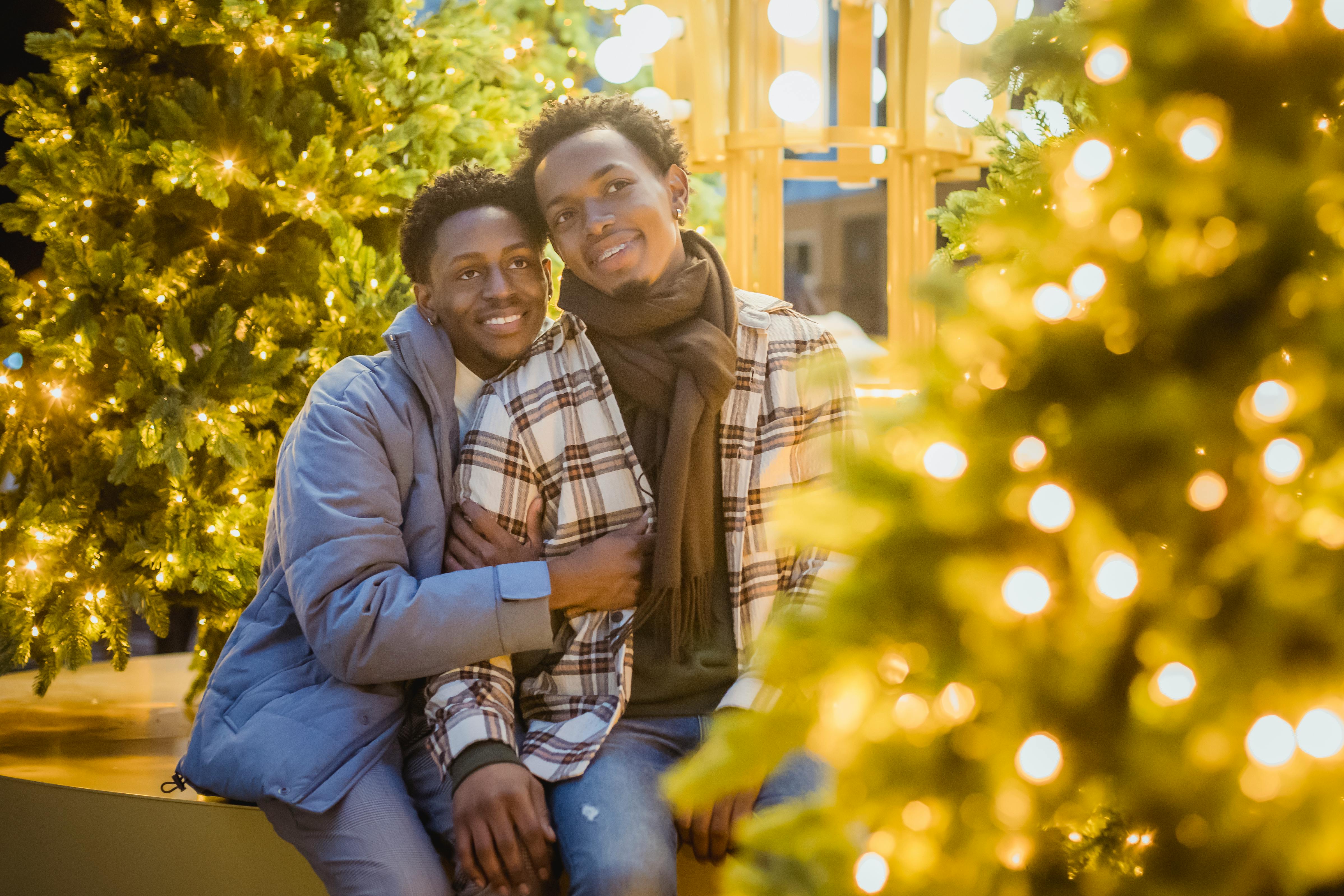 Positive African American homosexual couple cuddling and looking away while sitting on street near trees with glowing garlands at evening time