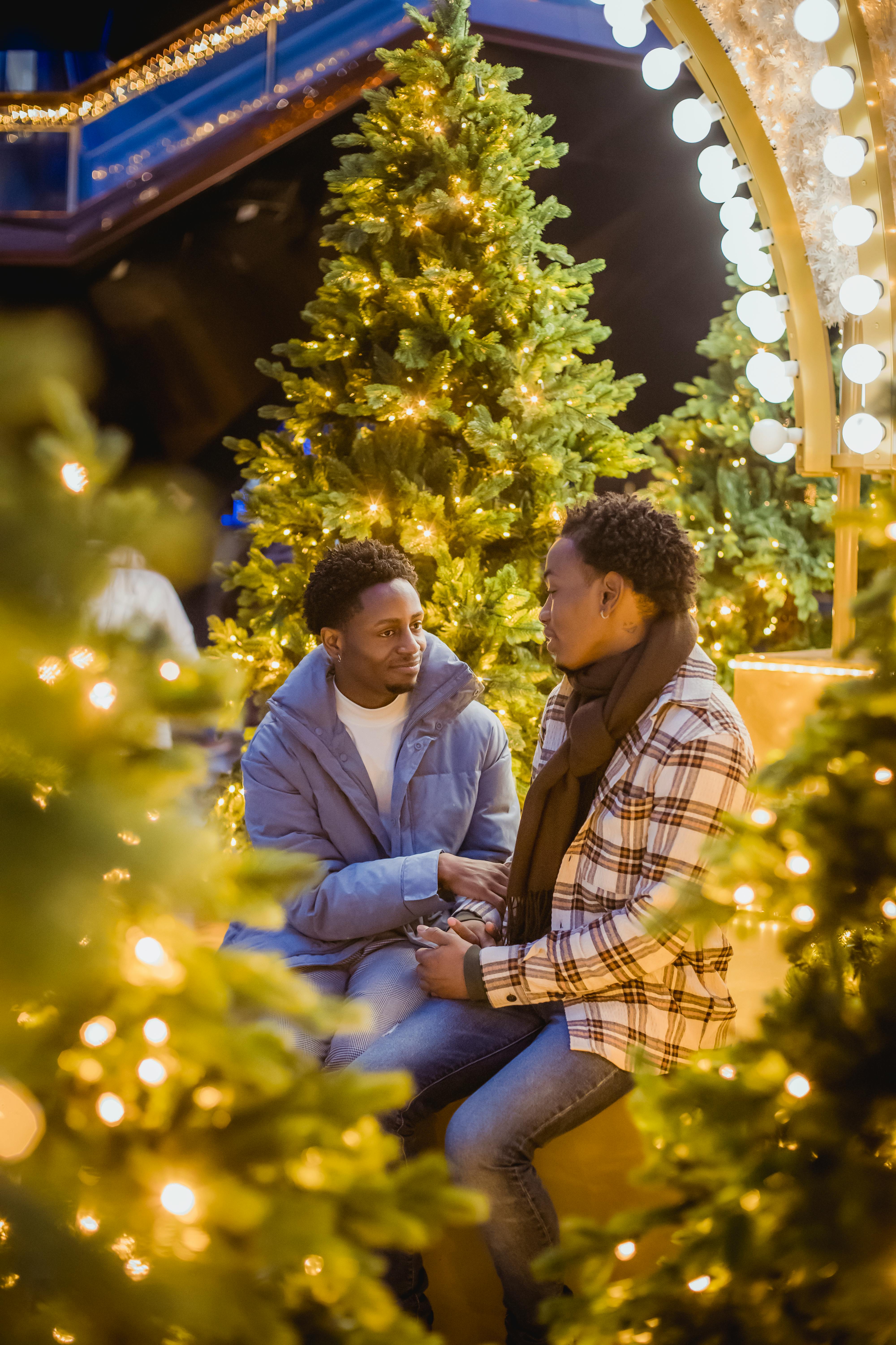Loving African American gays sitting amidst illuminated Christmas trees with garlands on street at evening time during romantic date on holidays