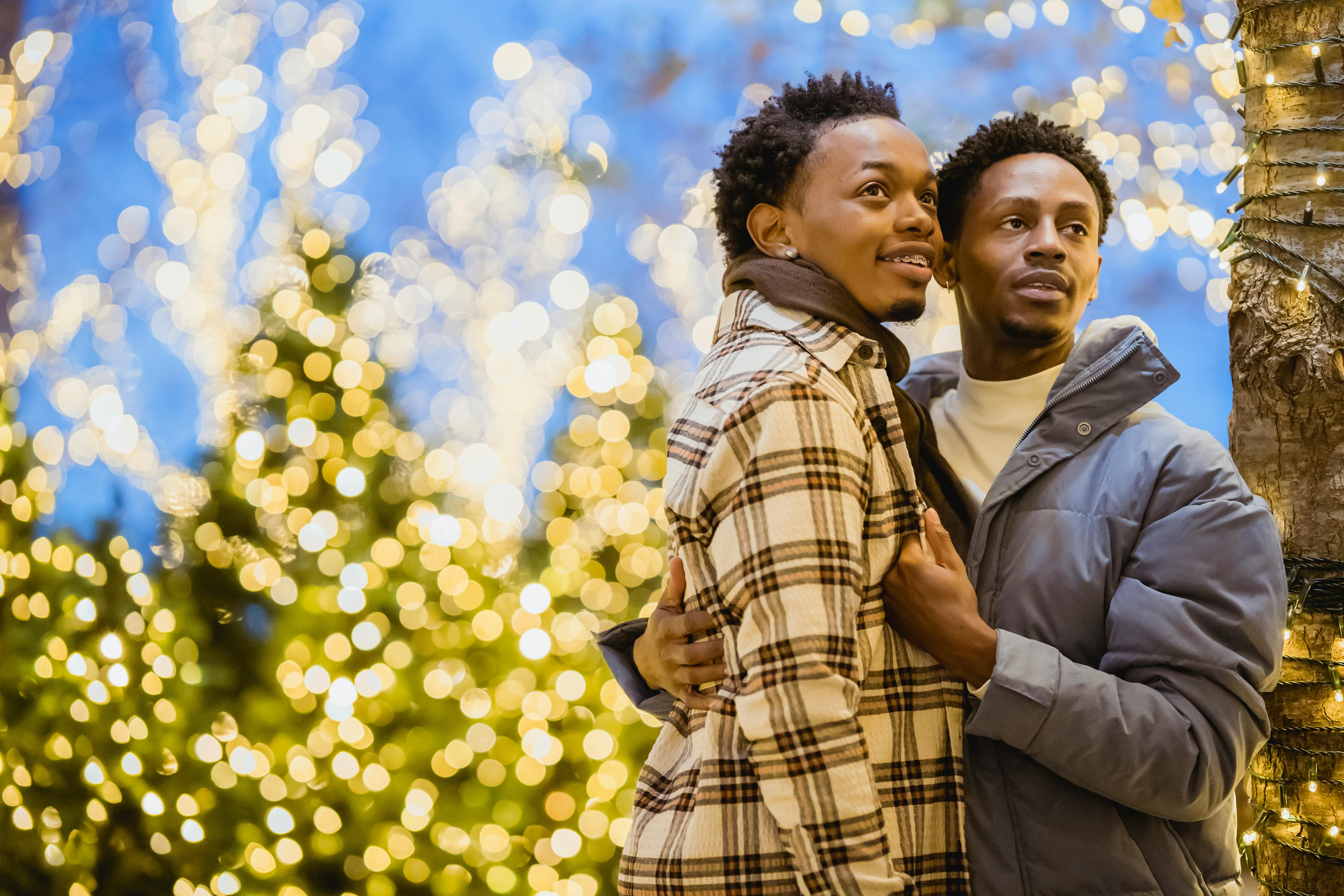 Positive African American homosexual couple cuddling and looking away while standing on street near illuminated Christmas trees on blurred background
