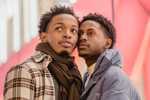 Curious African American homosexual couple in stylish wear touching faces and looking away while standing in city on blurred background