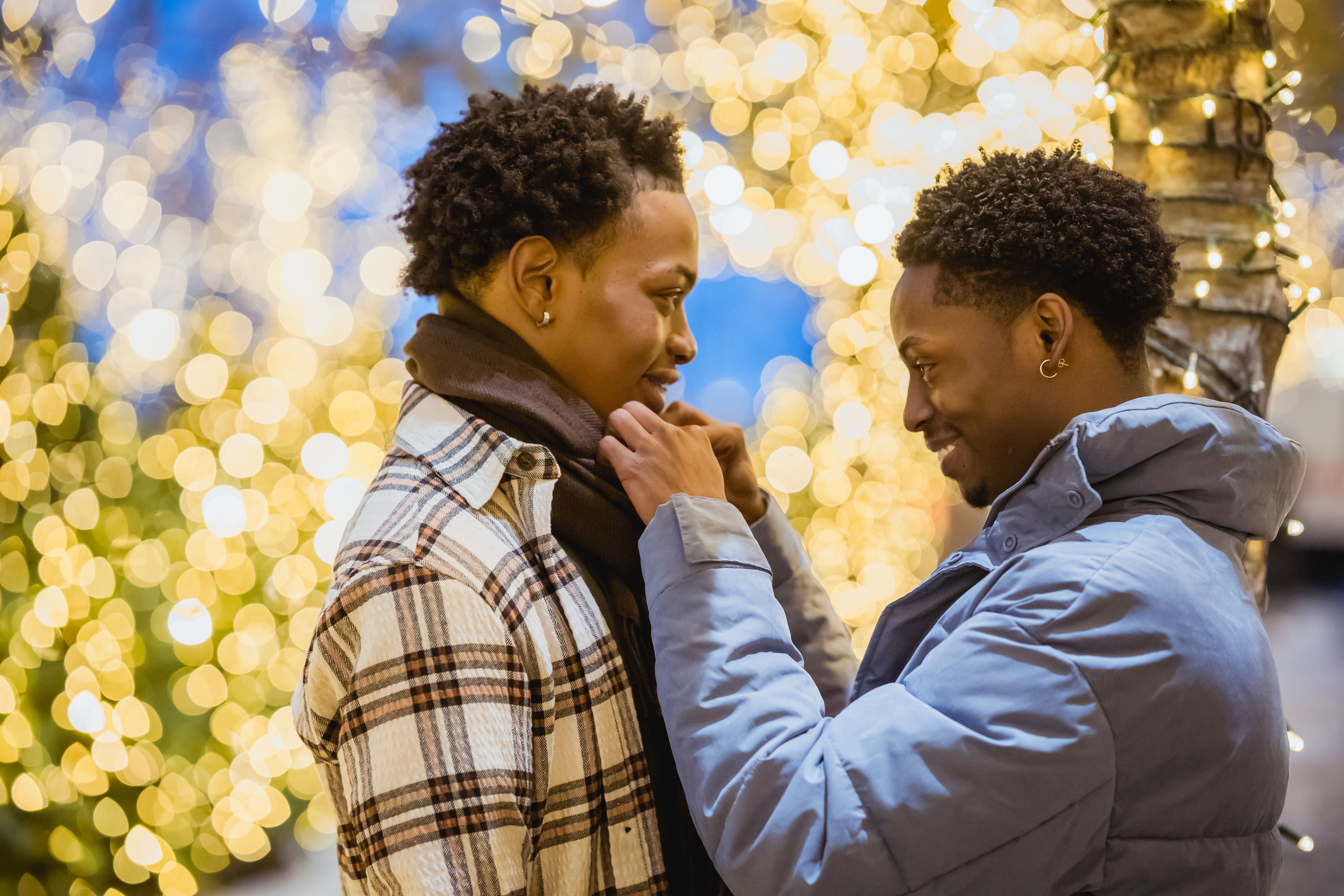 Side view of positive African American homosexual man straightening scarf on black partner while standing on street with glowing trees on blurred background
