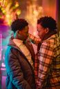 Side view of romantic African American gays looking at each other while standing near glass wall of building with colorful illumination