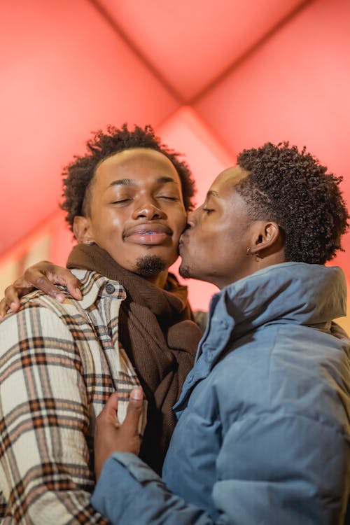 Tender African American homosexual man with closed eyes kissing cheerful black partner on cheek while standing on street during romantic date
