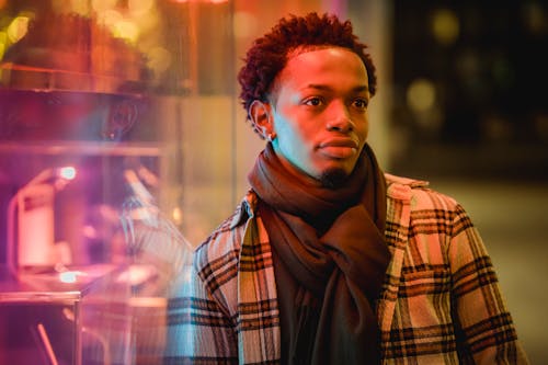 Stylish African American male in trendy outfit looking away thoughtfully while standing near glass wall of building with light at evening time