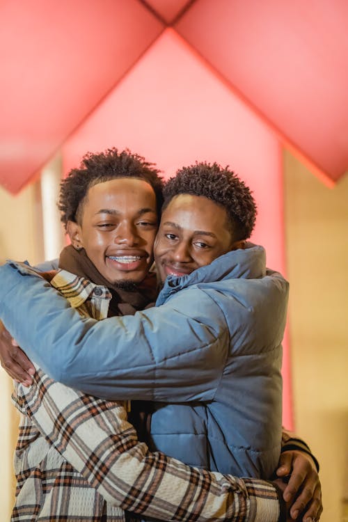 Delighted African American homosexual couple hugging and looking at camera while standing on street near wall of building in city