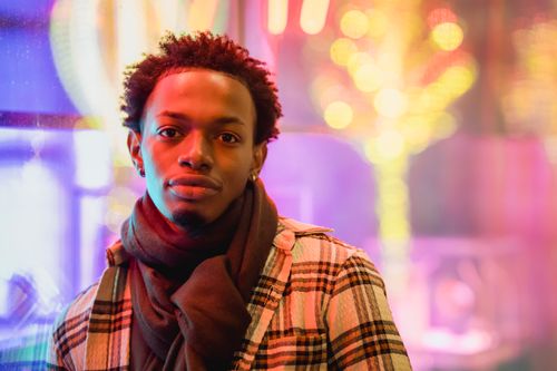 Free Content African American male wearing trendy outfit and scarf looking at camera while standing on street with illuminated lights at evening time on blurred background Stock Photo