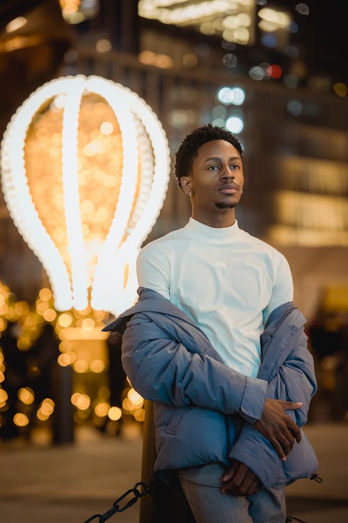 Confident African American male wearing jacket off shoulders looking away while standing on street with glowing lights on blurred background at evening time