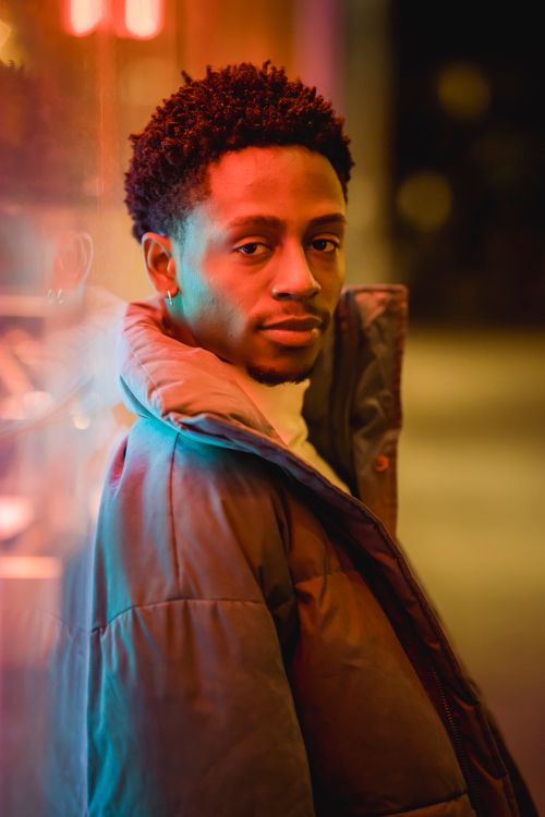 Free Side view of African American male in outerwear looking at camera near luminous building in evening Stock Photo