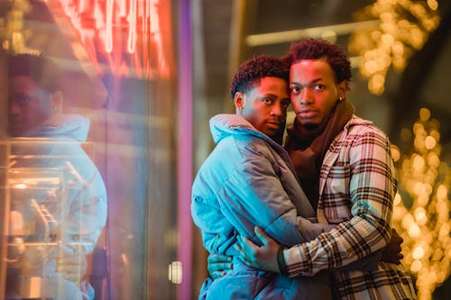 Happy African American couple of homosexual men hugging near window with neon glowing lights on blurred background of street