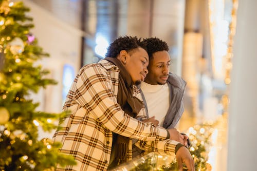 Positive African American couple of gays smiling and touching hands near Christmas tree with glowing garland